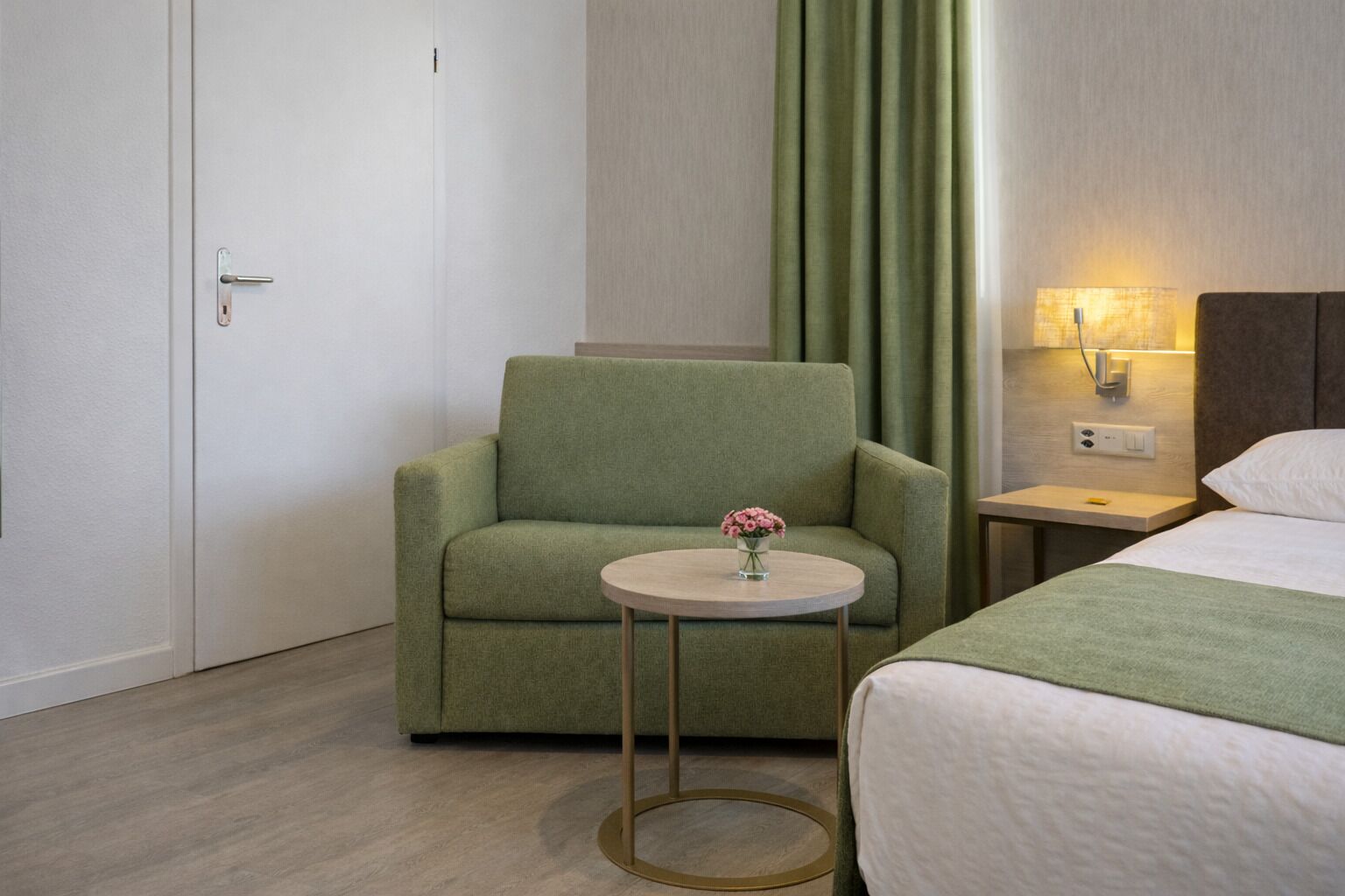 A hotel room with a green armchair, a small round table with a vase of flowers, a bed with a green throw, and a nightstand with a lamp.