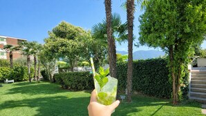Eine Hand hält ein Getränk mit Minzblättern, im Hintergrund ein gepflegter Rasen, Palmen und ein Hecke in sonniger Umgebung.
