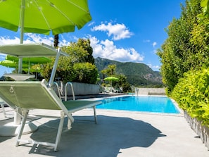 A pool area with lounge chairs and umbrellas, surrounded by greenery and mountains. - Hotel Nessi Locarno (Locarno)