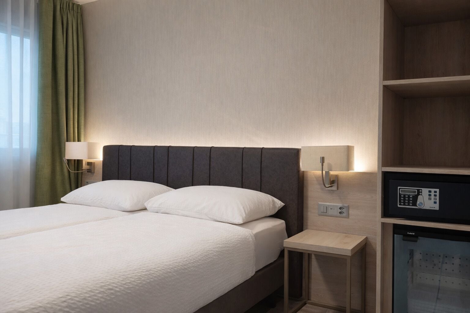 A hotel room with a bed, bedside lamps, a minibar, and a wooden shelf.