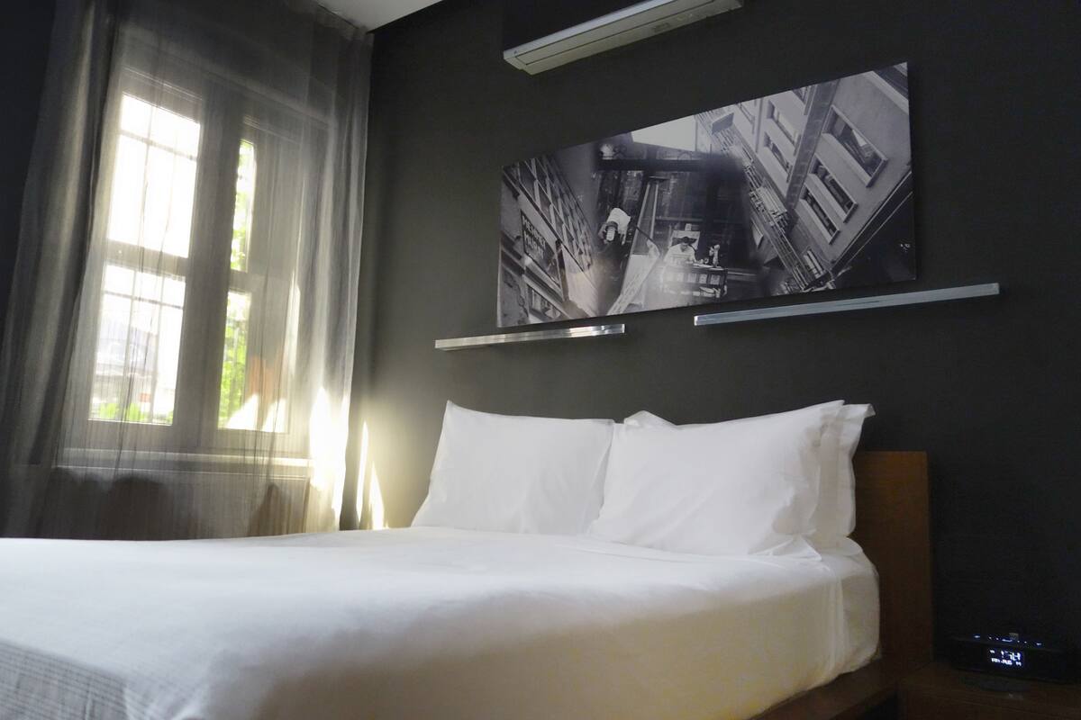 Habitación de hotel con una cama, un ventanal con cortinas translúcidas y una fotografía blanco y negro en la pared.