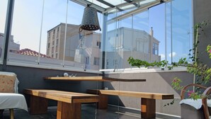 A modern outdoor seating area with wooden benches and a hanging light fixture, overlooking a cityscape through large windows. - I'zaz (Istanbul)