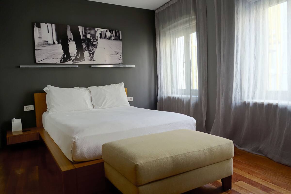 Un dormitorio con cama, mesita de noche, cortinas translúcidas en la ventana y una obra de arte colgada en la pared.