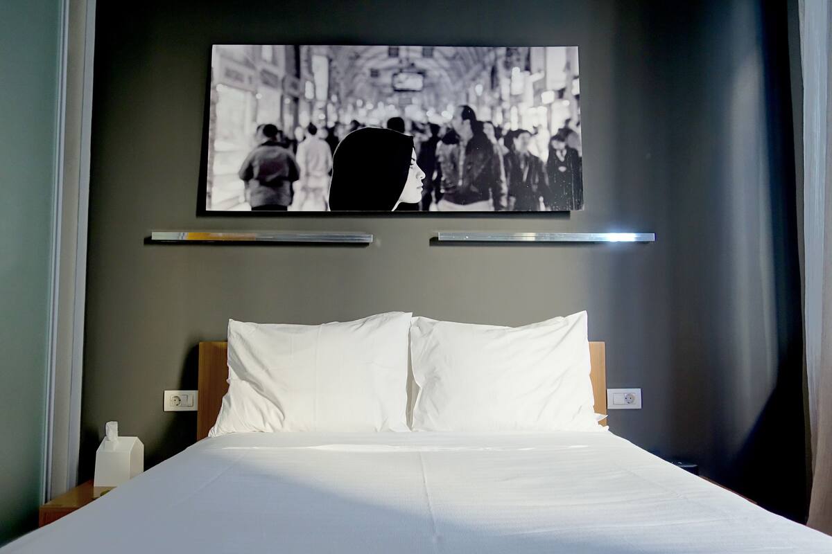Habitación de hotel con cama, almohadas, mesita de noche, caja de pañuelos y una fotografía en blanco y negro enmarcada en la pared.