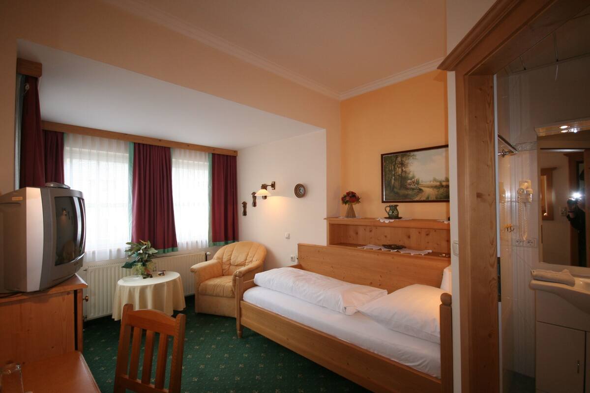 Ein gemütliches Hotelzimmer mit einem Einzelbett, einem Sessel, einem kleinen Tisch, einem Fernseher und einem Bild an der Wand.