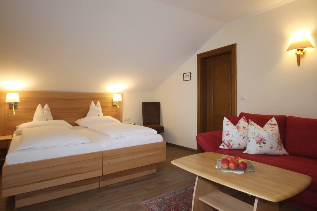 Ein Schlafzimmer mit einem hölzernen Bett, einem roten Schlafsofa und einem kleinen Tisch mit Obst.