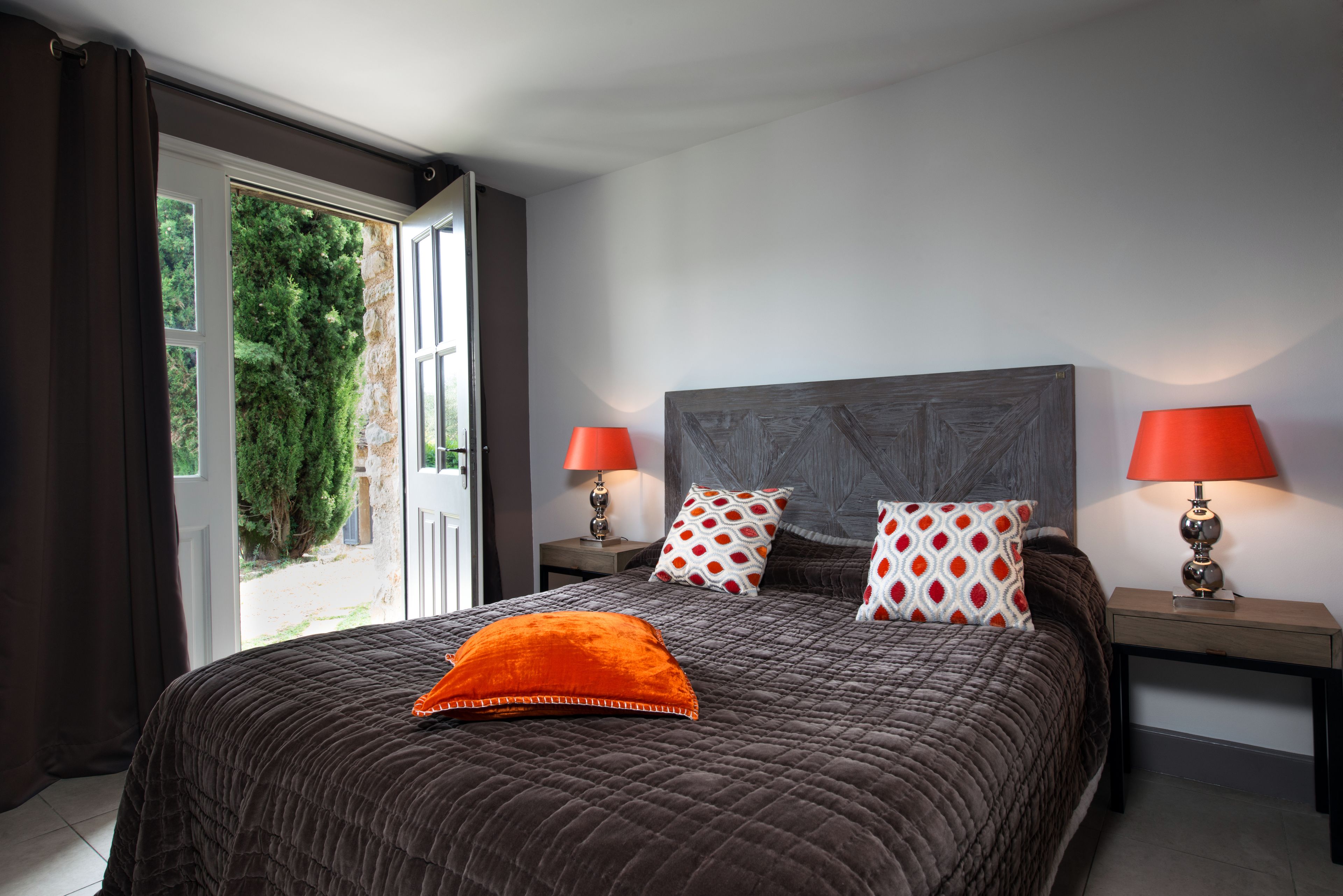 Une chambre avec un grand lit, deux tables de chevet, deux lampes rouges et une vue sur la verdure par une porte ouverte.