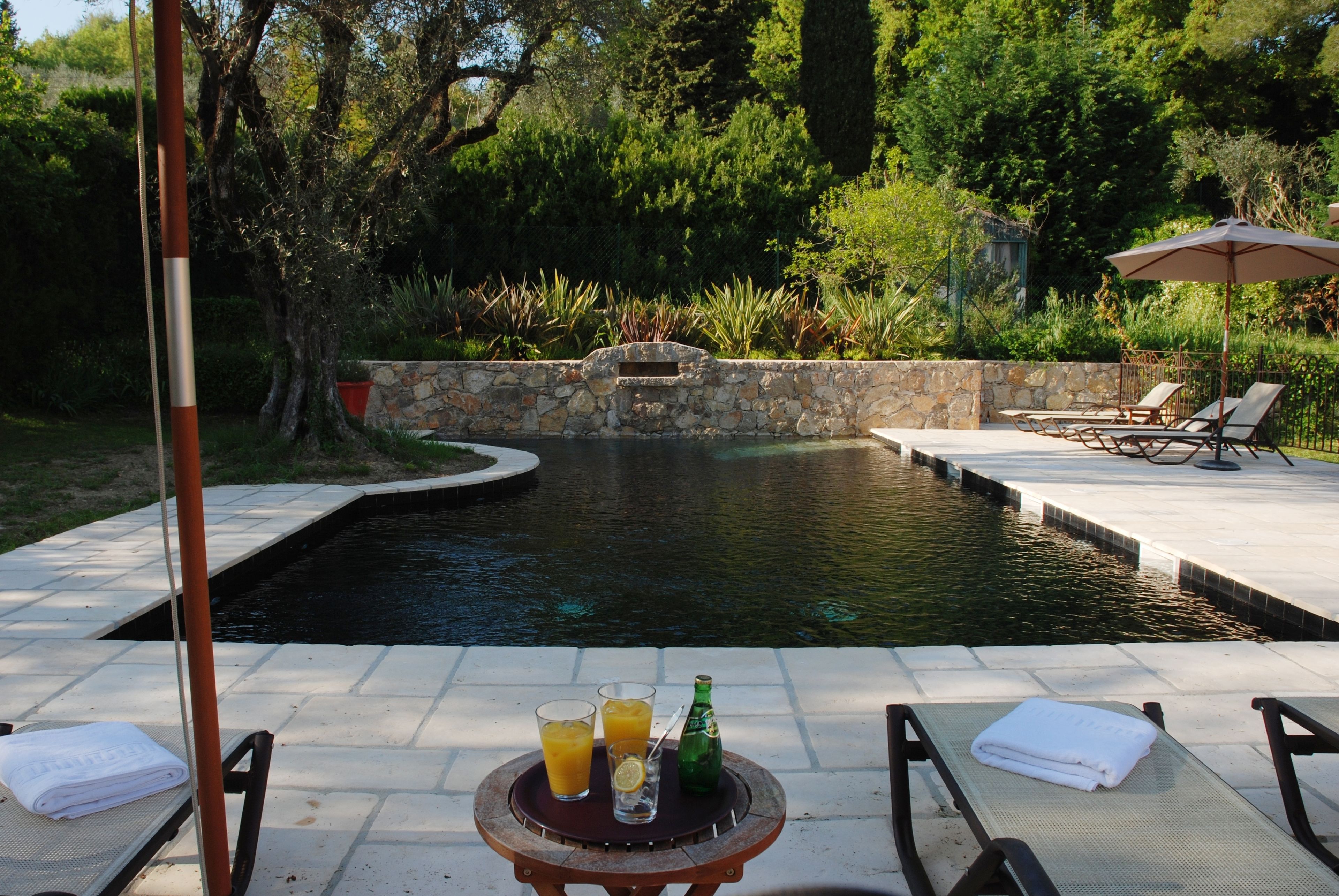 Una piscina con pared de piedra, sillas de descanso y una mesa con bebidas y una toalla.