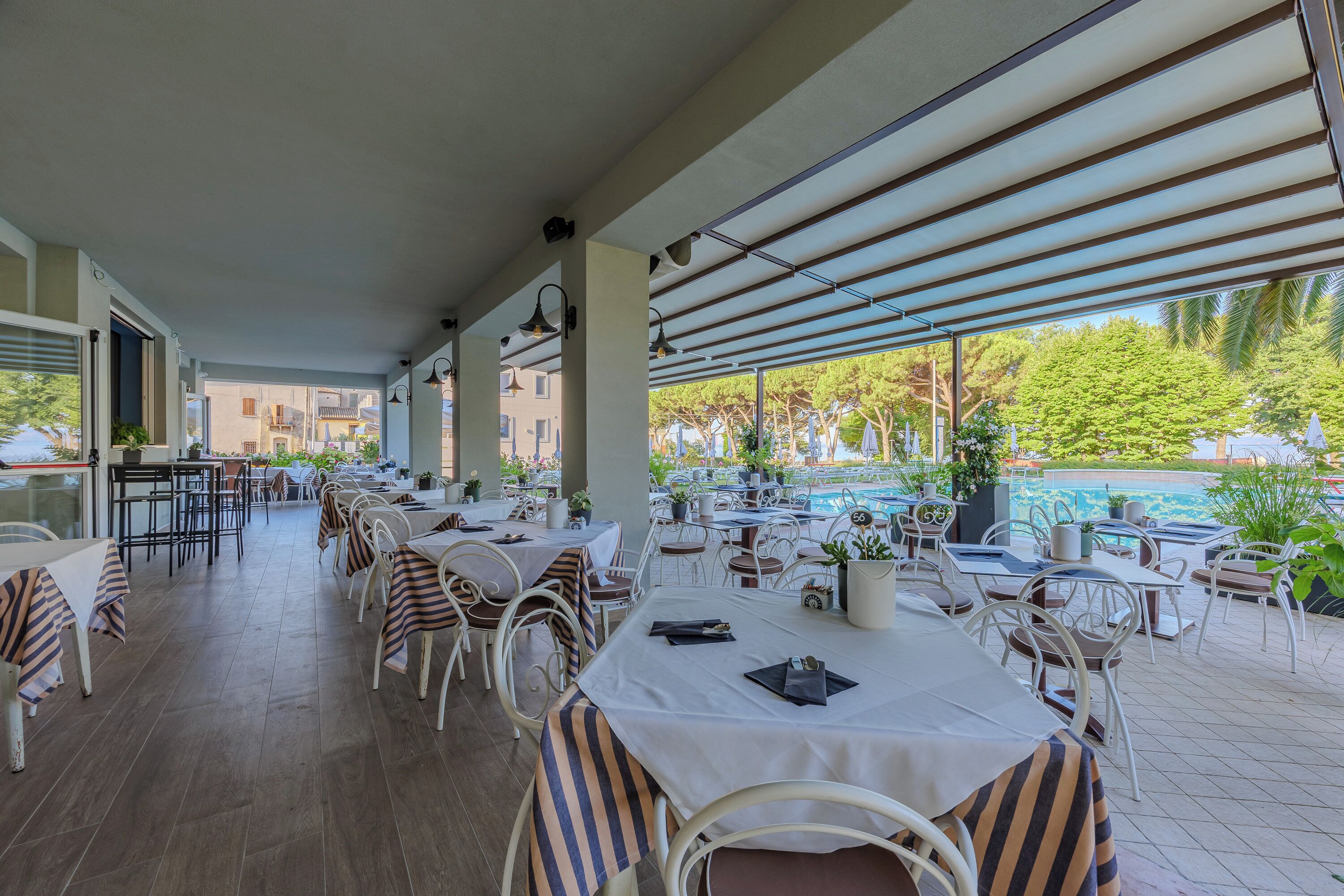 Un área de comedor al aire libre techada con mesas y sillas, una piscina al fondo y vistas a edificios y árboles.