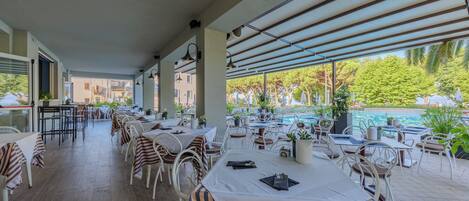 Un área de comedor al aire libre techada con mesas y sillas, una piscina al fondo y vistas a edificios y árboles.