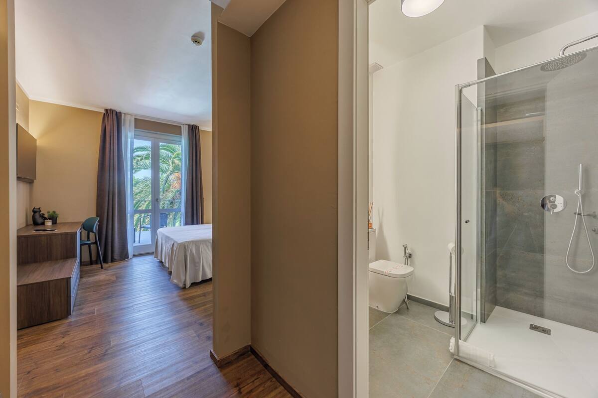 A hotel room with a wooden floor, a bed, a desk, a chair, a shower with a glass door, and a view of palm trees through the window.