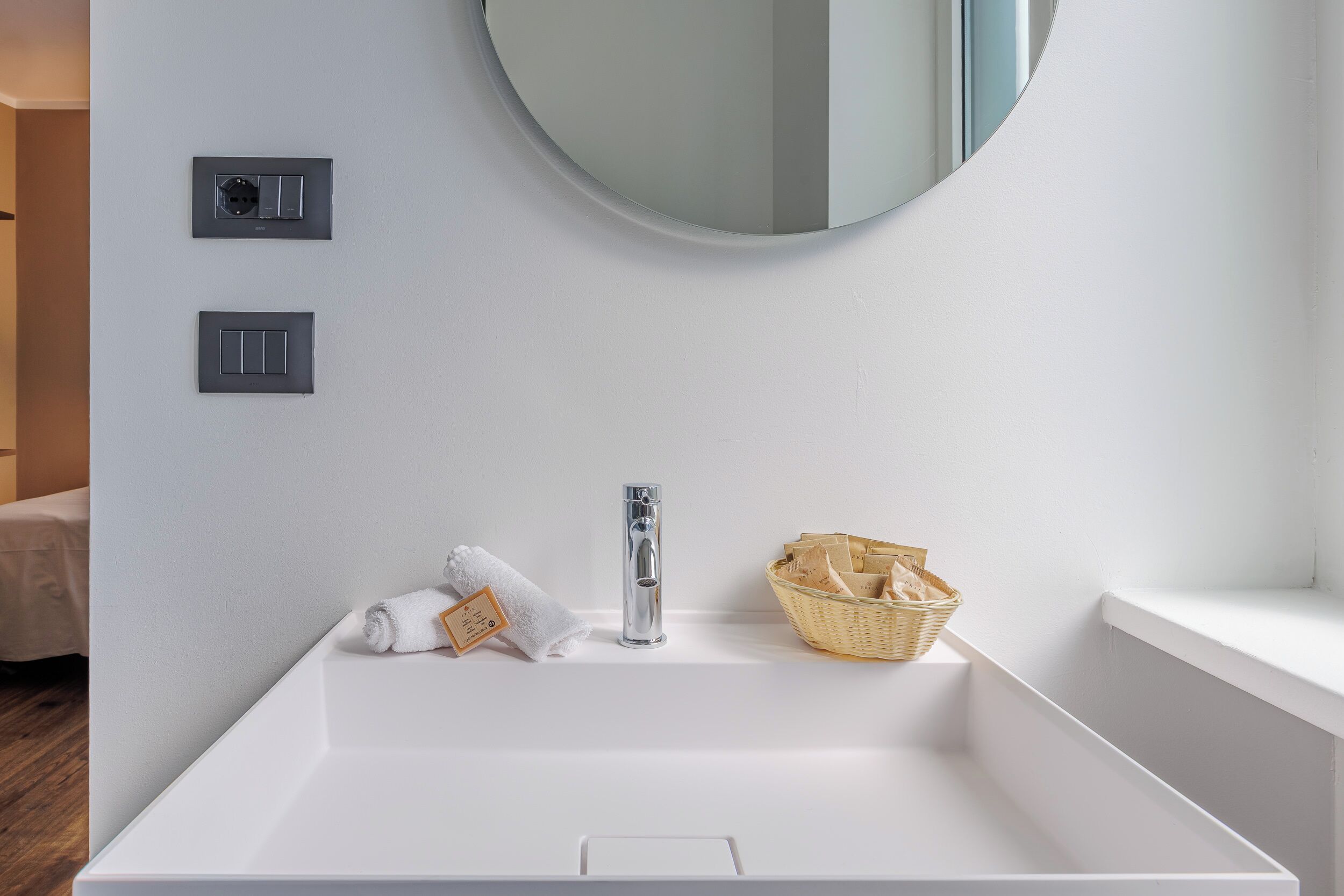 A bathroom with a large white sink, a round mirror, and two electrical outlets on the wall.
