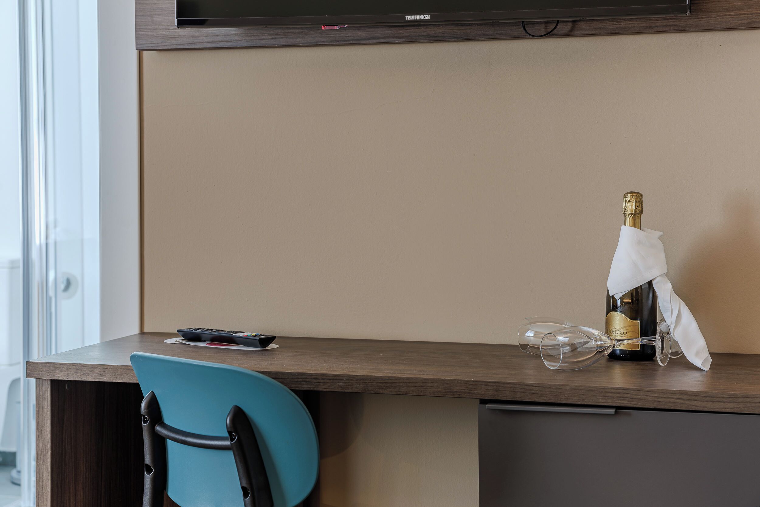 Een hotelkamer met een houten bureau, een blauwe stoel, een televisie en een fles champagne met een glas.
