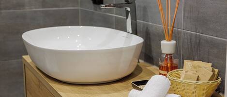 Un baño con un lavamanos blanco de diseño, un grifo moderno, un difusor de cañas, una cesta de mimbre con artículos de aseo y una toalla blanca doblada sobre un mueble de madera.
