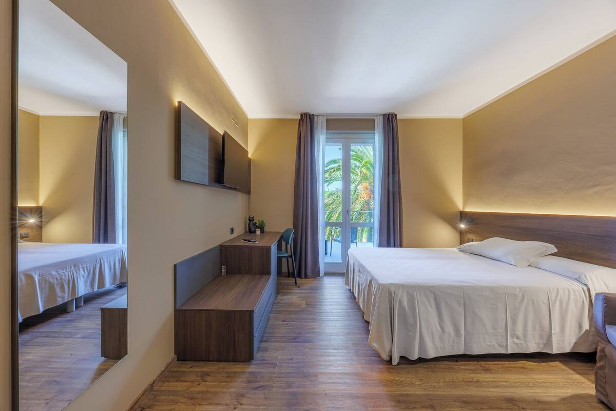 A modern hotel room with a large bed, a desk, and a balcony with a view of palm trees.