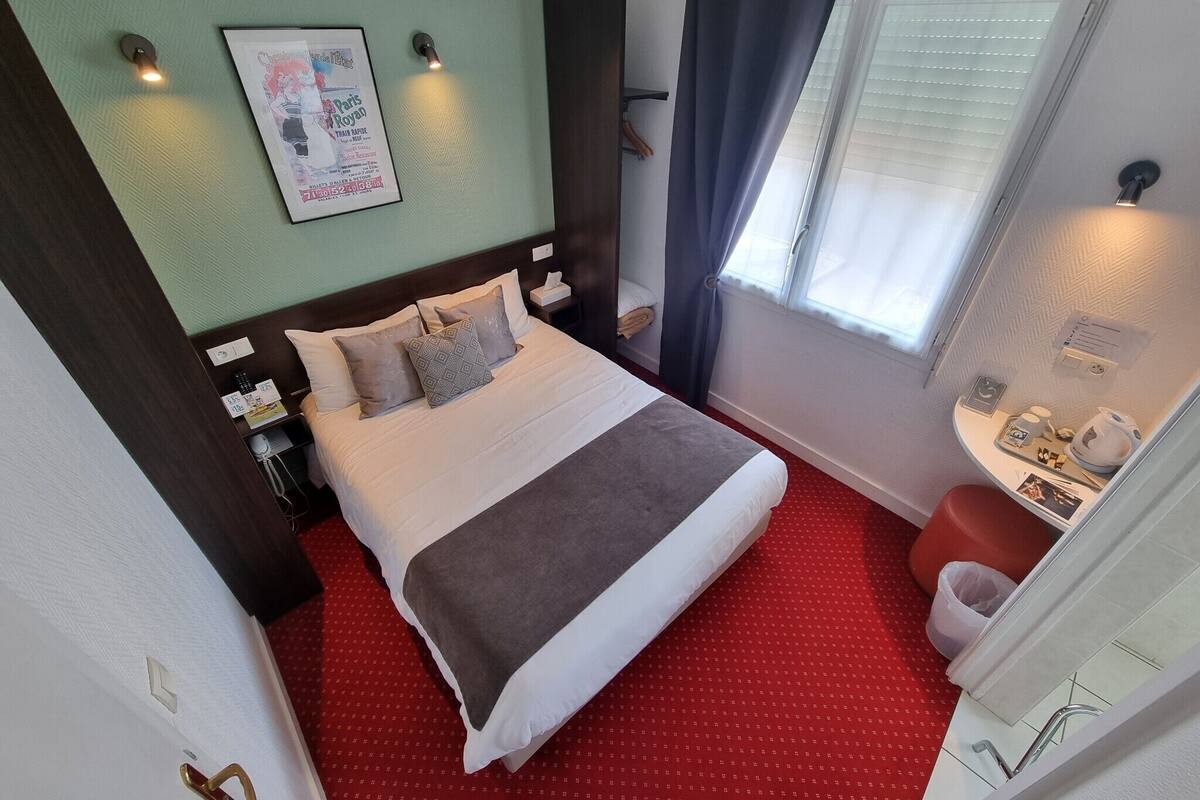 Habitación de hotel con cama, mesita de noche, lámpara, ventana con cortinas y una pequeña mesa con tetera y tazas.