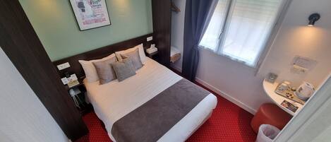 A hotel room with a bed, bedside table, lamp, window with curtains, and a small table with a kettle and cups.