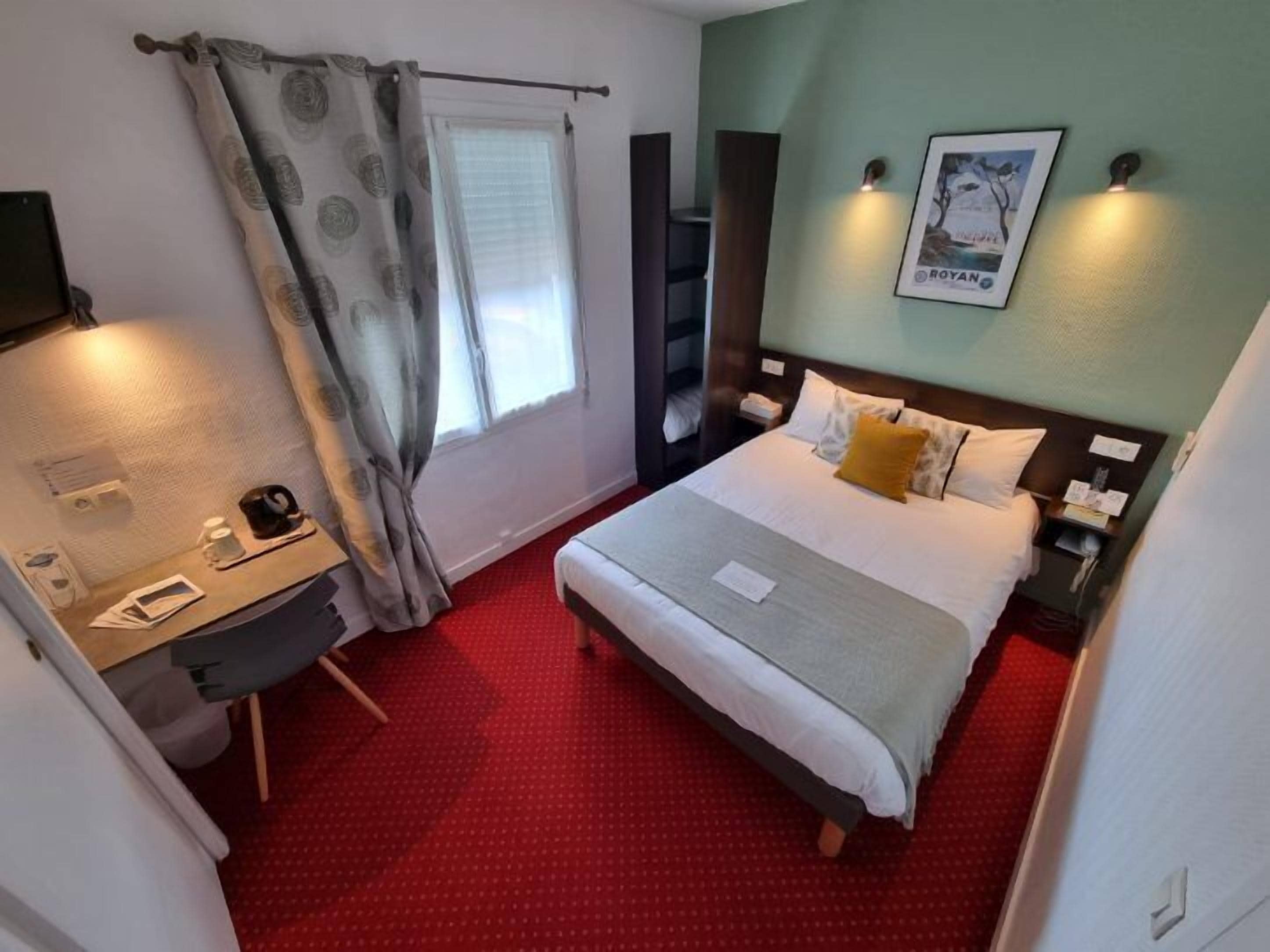 Habitació d'hotel amb llit, escriptori, cadira, taula petita, taulell de nit, làmpada, quadre a la paret i finestra amb cortines.