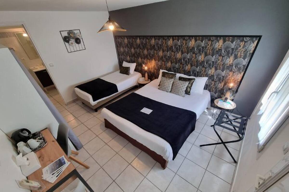 Habitación de hotel moderna con dos camas individuales, una cama grande, una mesita con una silla y un cabecero decorativo.
