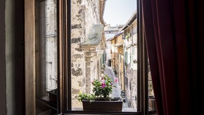 Uma janela com vista para uma rua estreita, ladeada por edifícios antigos, um vaso de planta no parapeito e uma cortina ligeiramente afastada.