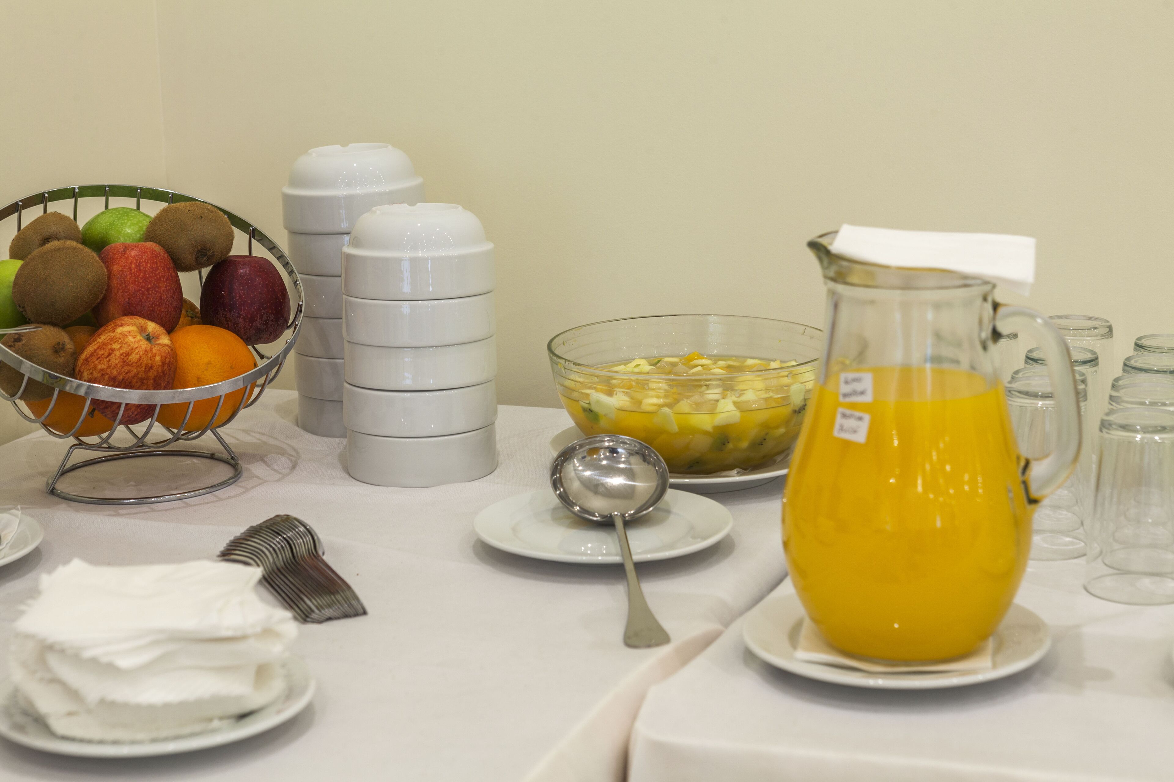 A table with a fruit basket, a pitcher of orange juice, a bowl of cereal, and several white cups.