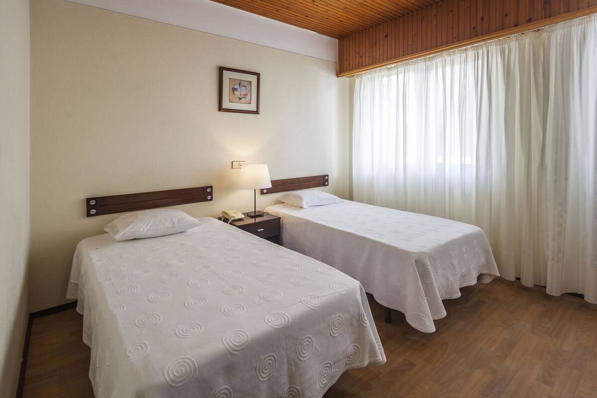 A hotel room with two single beds, a nightstand with a lamp, a telephone, and a framed picture on the wall.