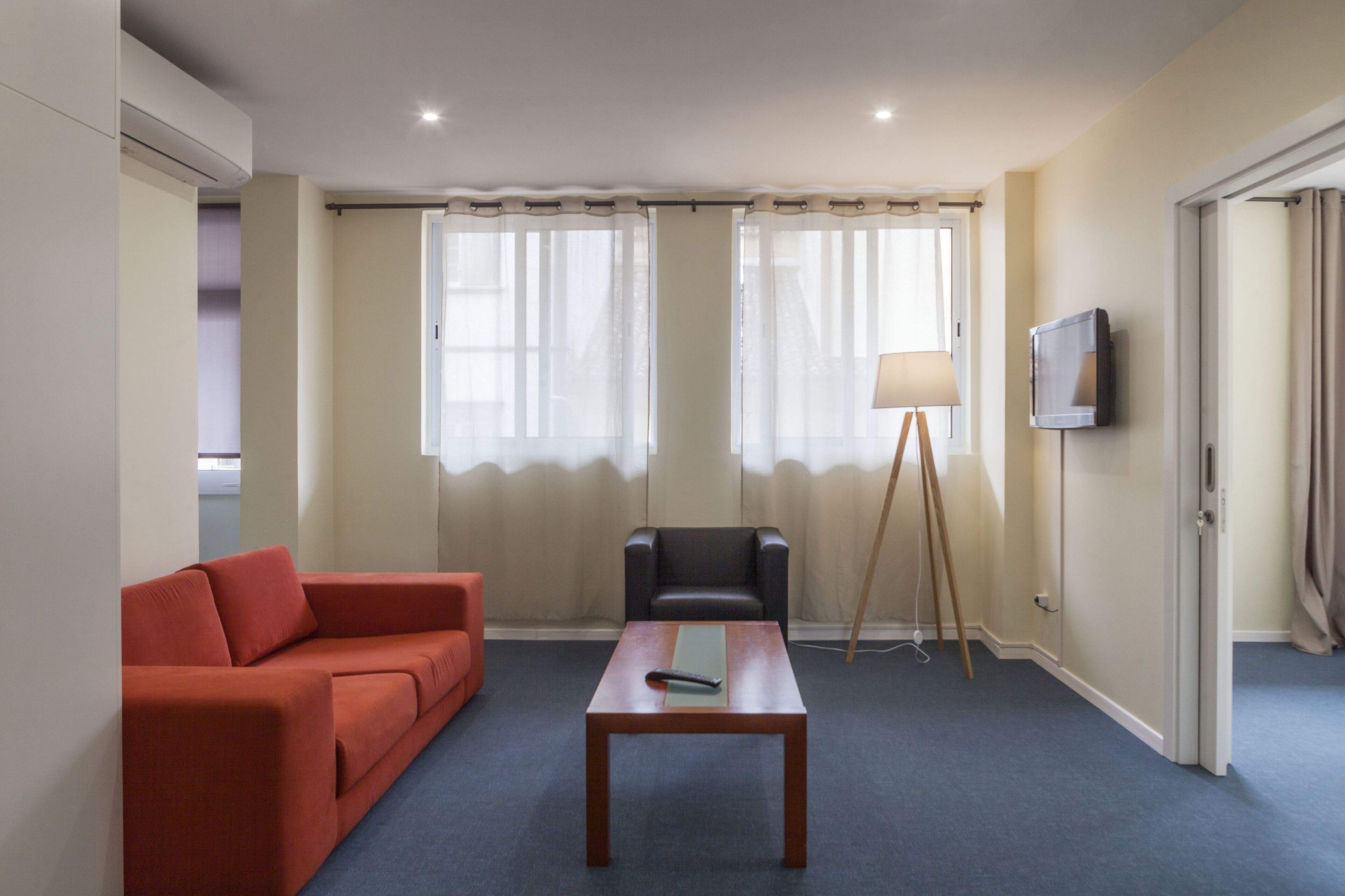 A hotel room with a sofa, a table, a chair, a floor lamp, and a TV mounted on the wall.