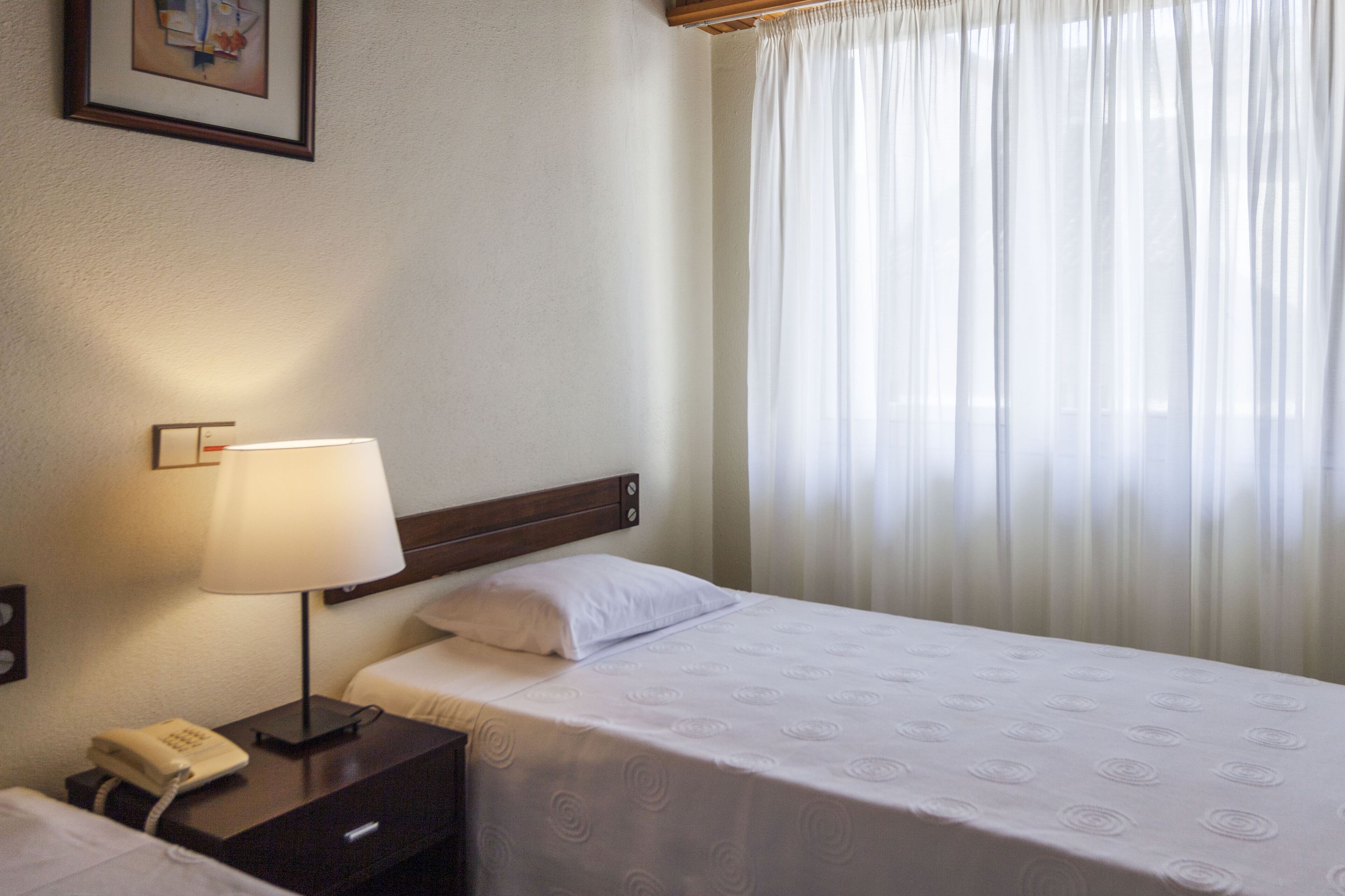 A hotel room with a bed, a bedside lamp, a telephone, and a window with sheer curtains.