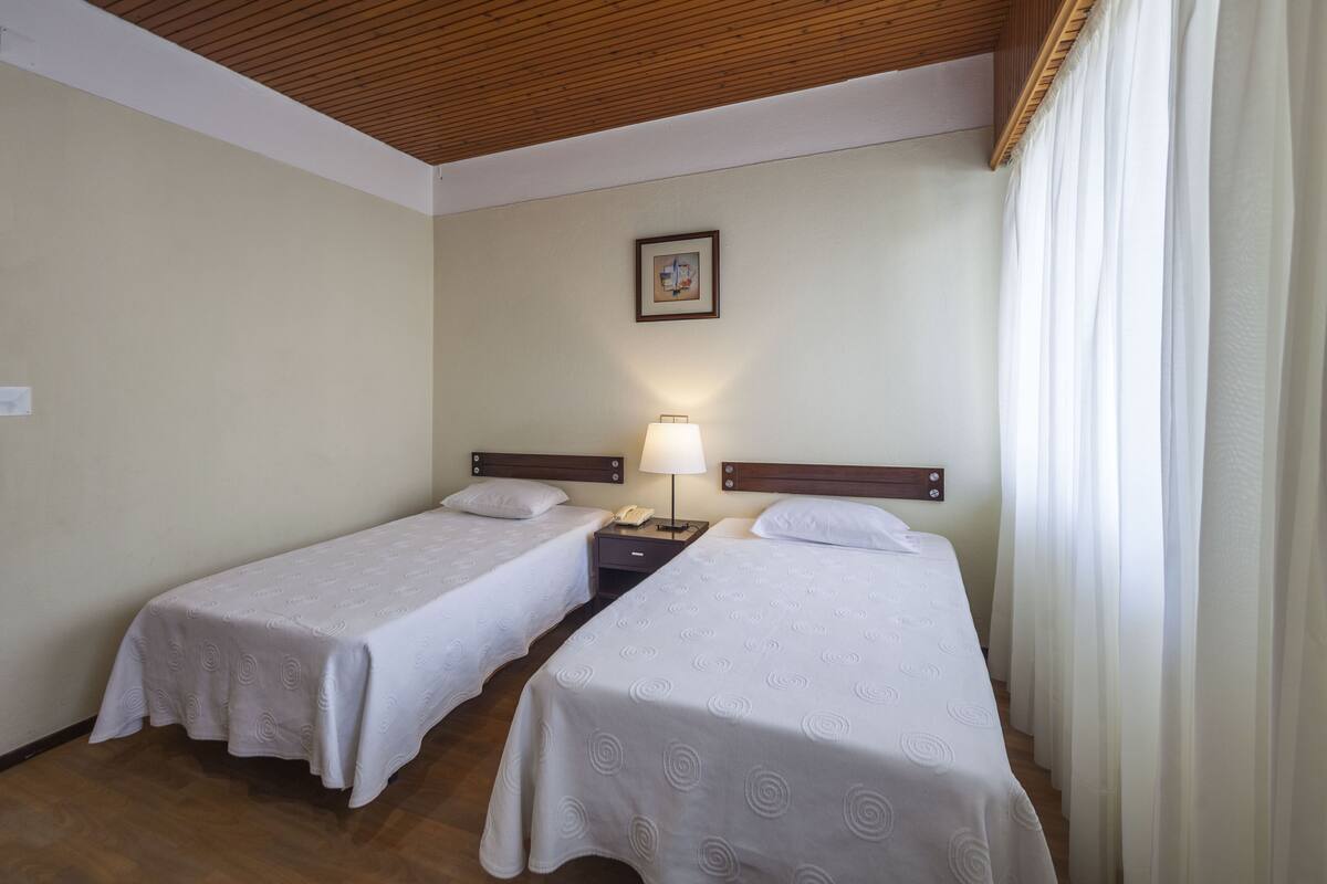A hotel room with two single beds, a nightstand with a lamp, a framed picture on the wall, and a window with curtains.