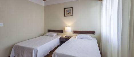 A hotel room with two single beds, a nightstand with a lamp, a framed picture on the wall, and a window with curtains.