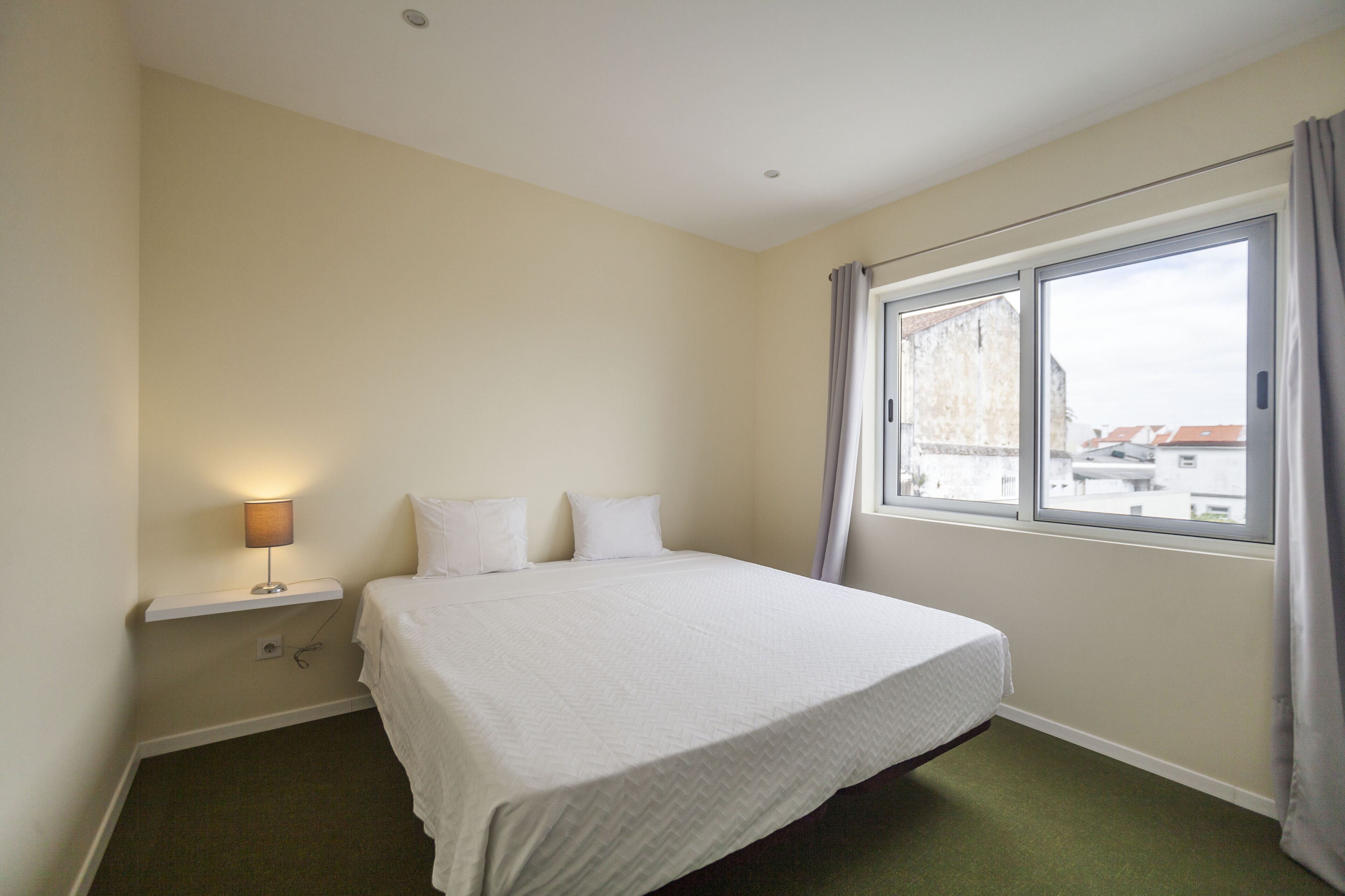 Une chambre d’hôtel avec un lit simple, une table de chevet avec une lampe, une fenêtre avec des rideaux et une vue sur les bâtiments environnants.