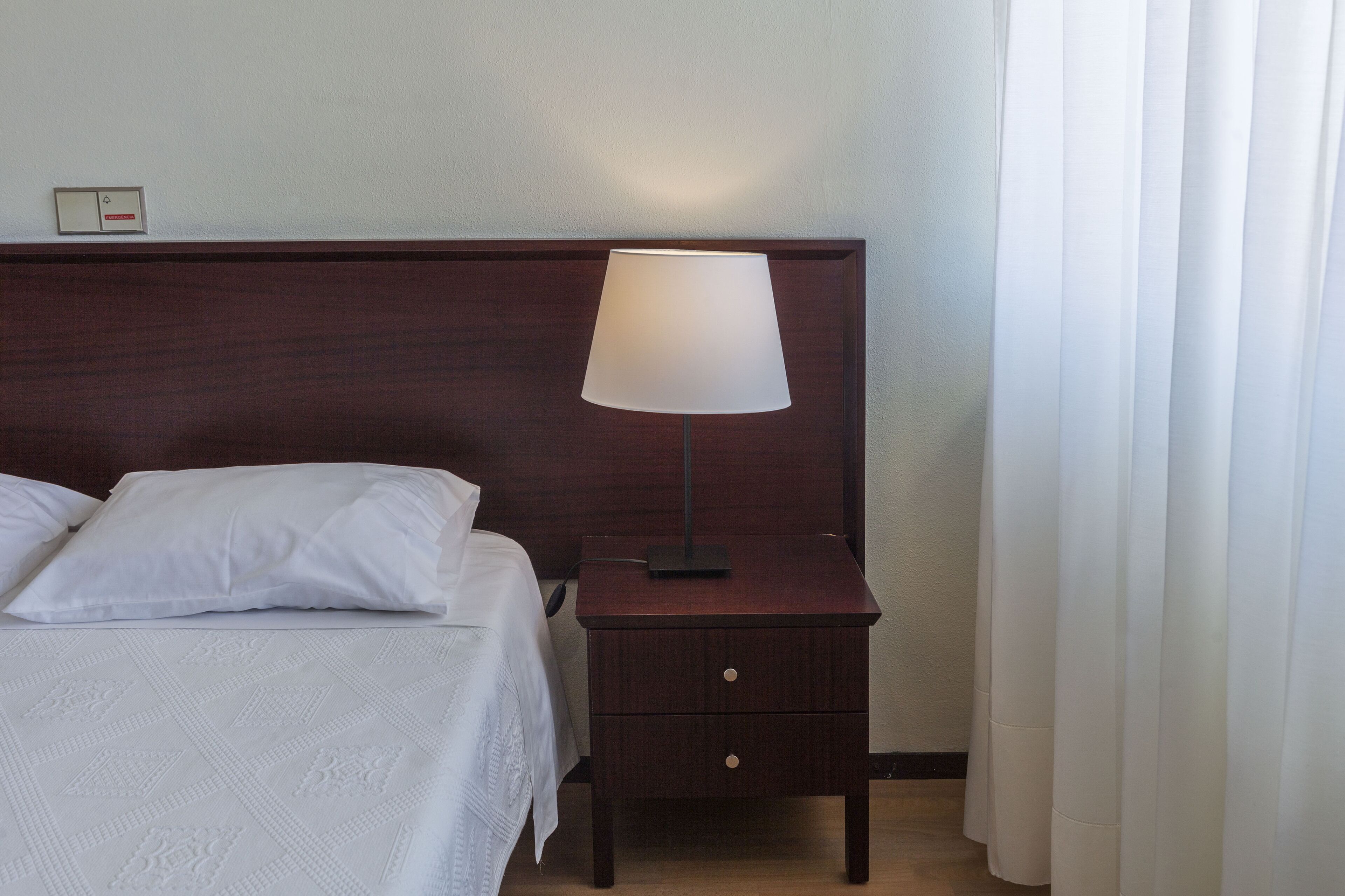 A neatly made bed with white linens, a wooden nightstand with a lamp, and a wall-mounted light switch.