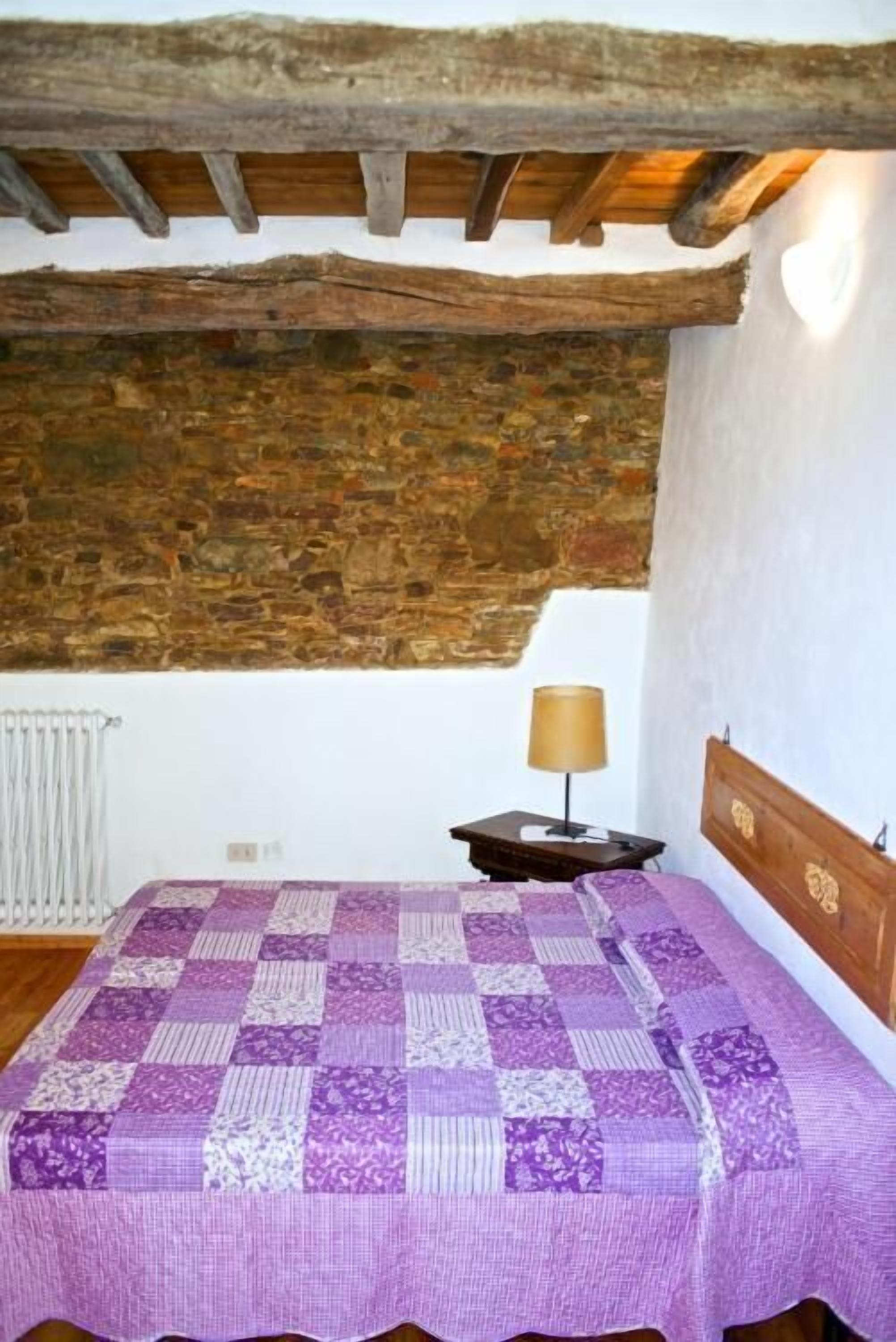 A bedroom with a wooden ceiling, a stone wall, a bed with a purple quilt, a nightstand with a lamp, and a radiator.