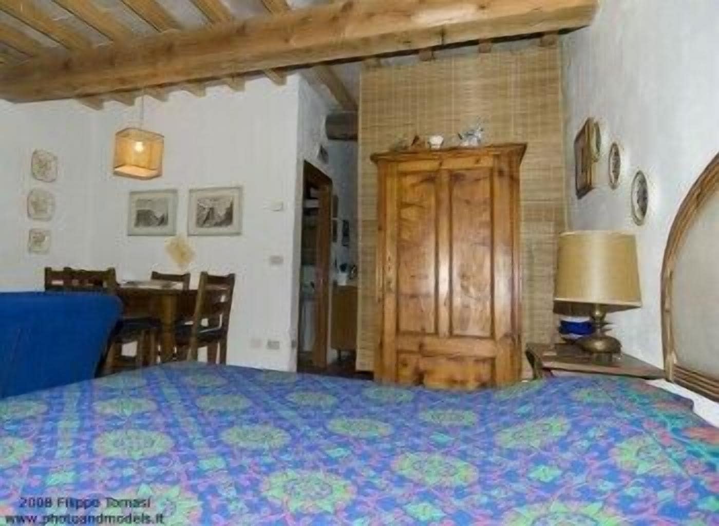A bedroom with a bed, a wooden wardrobe, a desk with chairs, and a lamp.