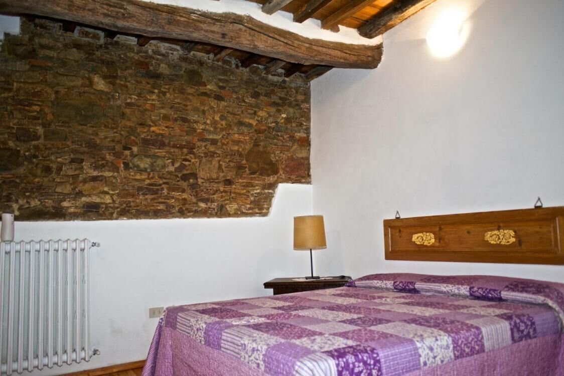 A bedroom with a stone wall, a wooden ceiling, a bed with a patterned bedspread, a bedside table with a lamp, and a radiator.