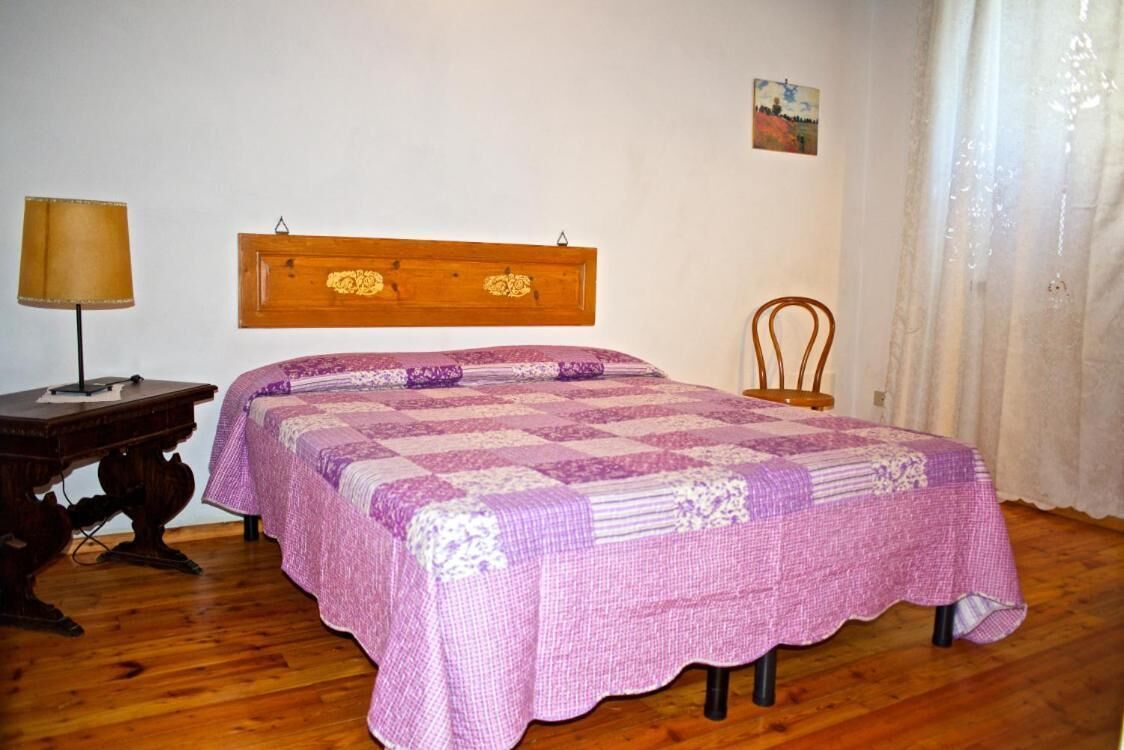Un dormitorio con cabecera de madera, una cama con edredón blanco y morado a cuadros, una mesita de noche de madera con lámpara y una pequeña silla.