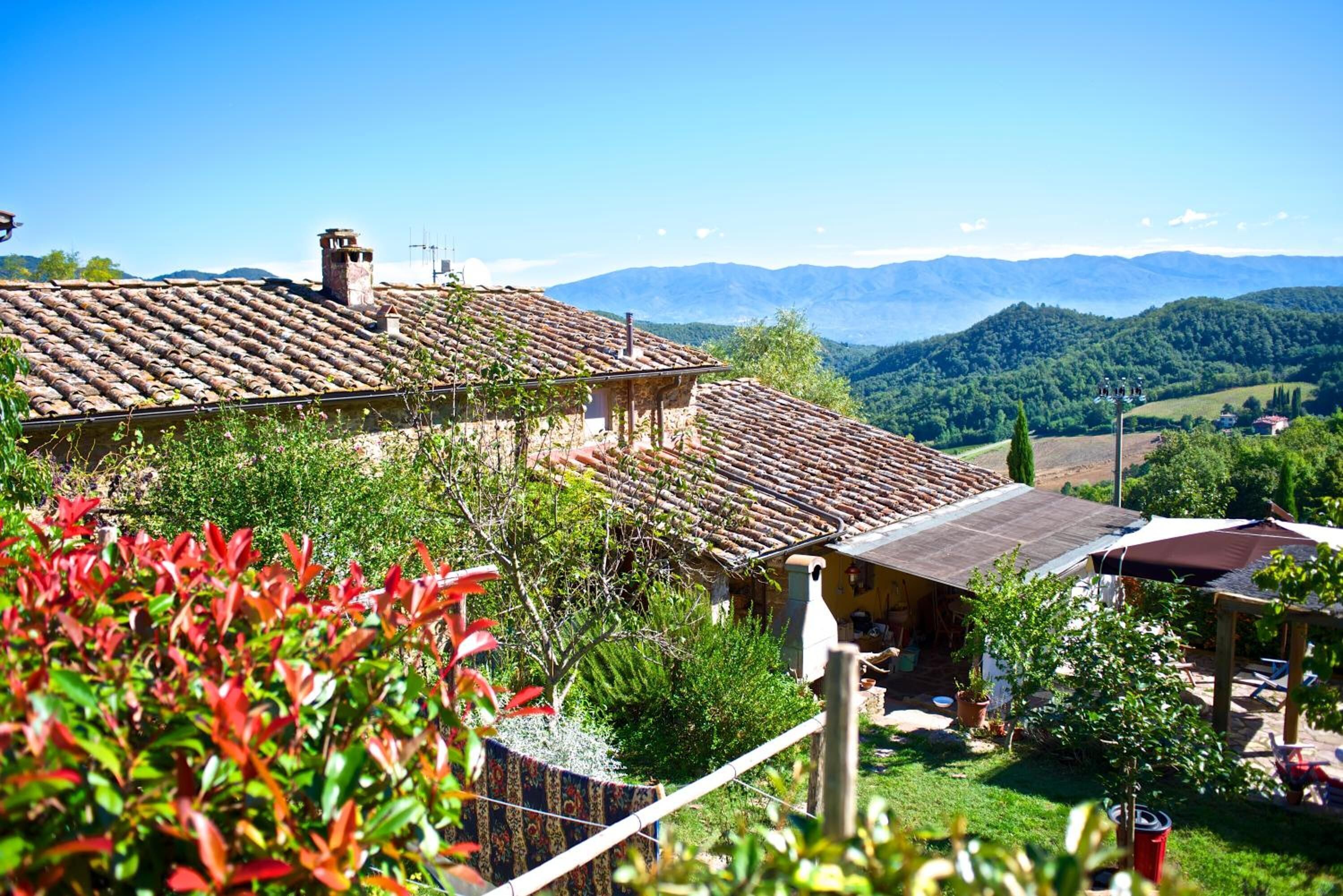 A rustic building with a tiled roof, surrounded by greenery and mountains in the background.