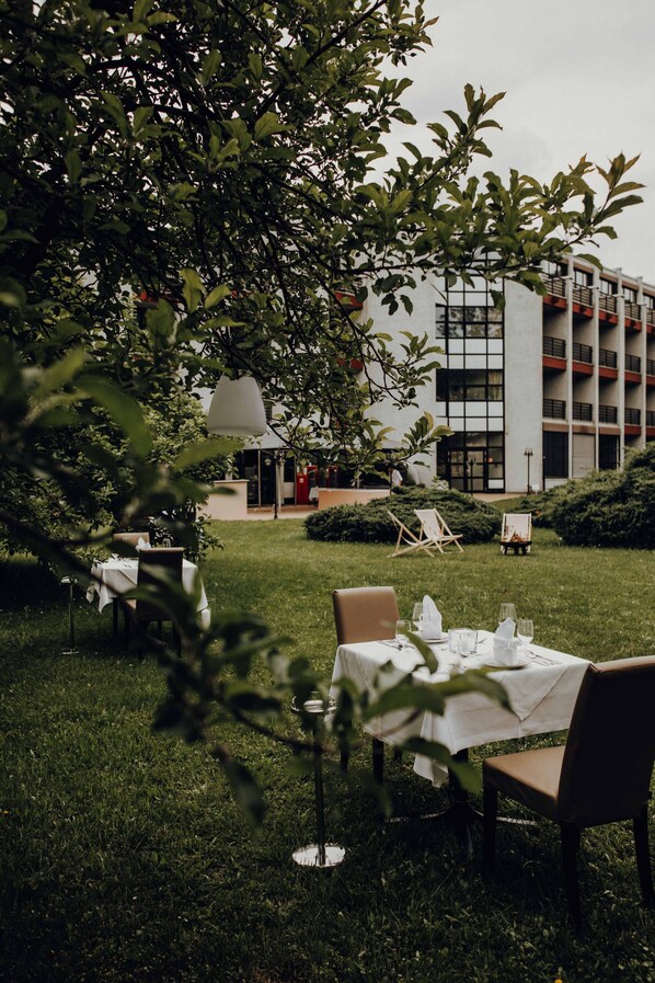 An outdoor dining area with tables set for two, surrounded by greenery and a building in the background. - Parkhotel Brunauer (Salzburg)