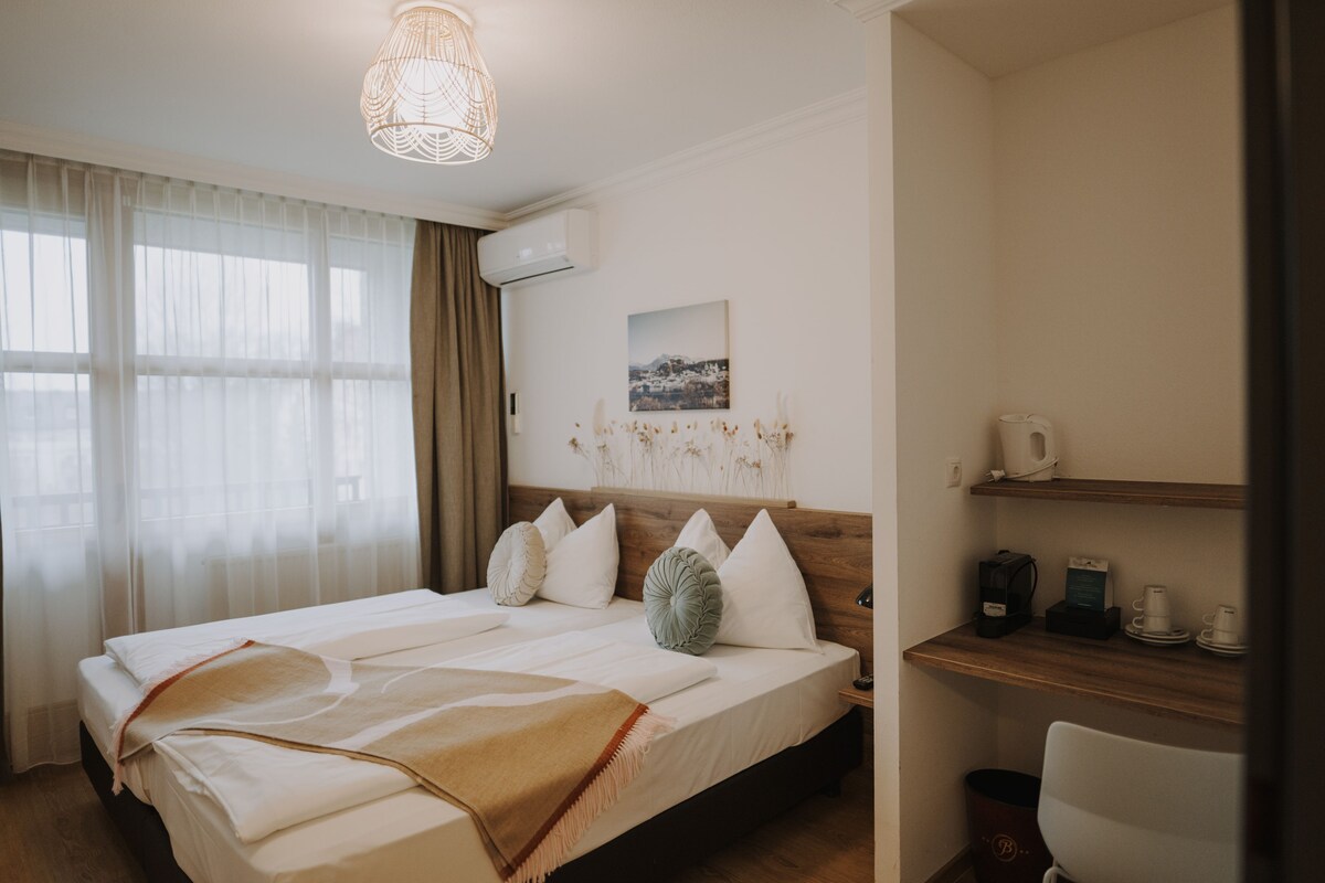 Una habitación de hotel con una cama, una mesita de noche con una lámpara, un estante con tazas y una bolsa, y un cuadro en la pared.