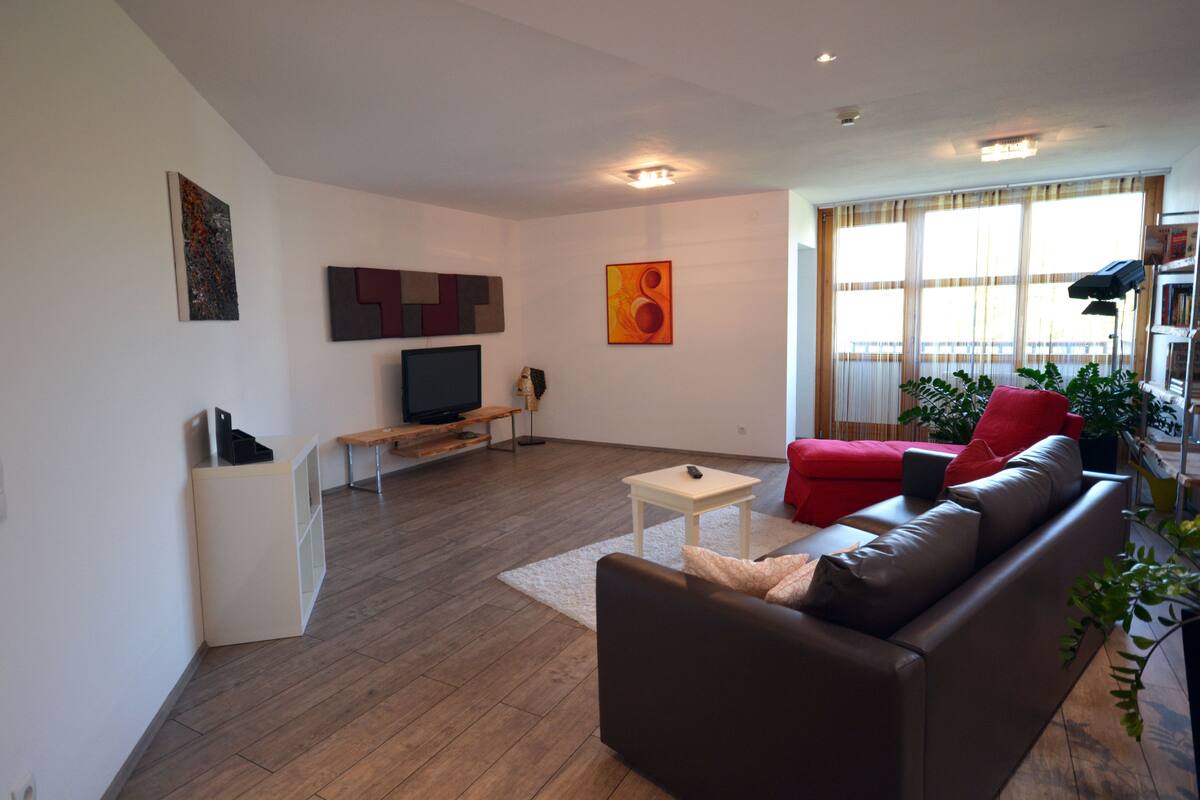 Una sala de estar moderna con un sofá de cuero marrón, un sillón rojo, una mesa de centro blanca y un televisor de pantalla plana sobre un soporte.