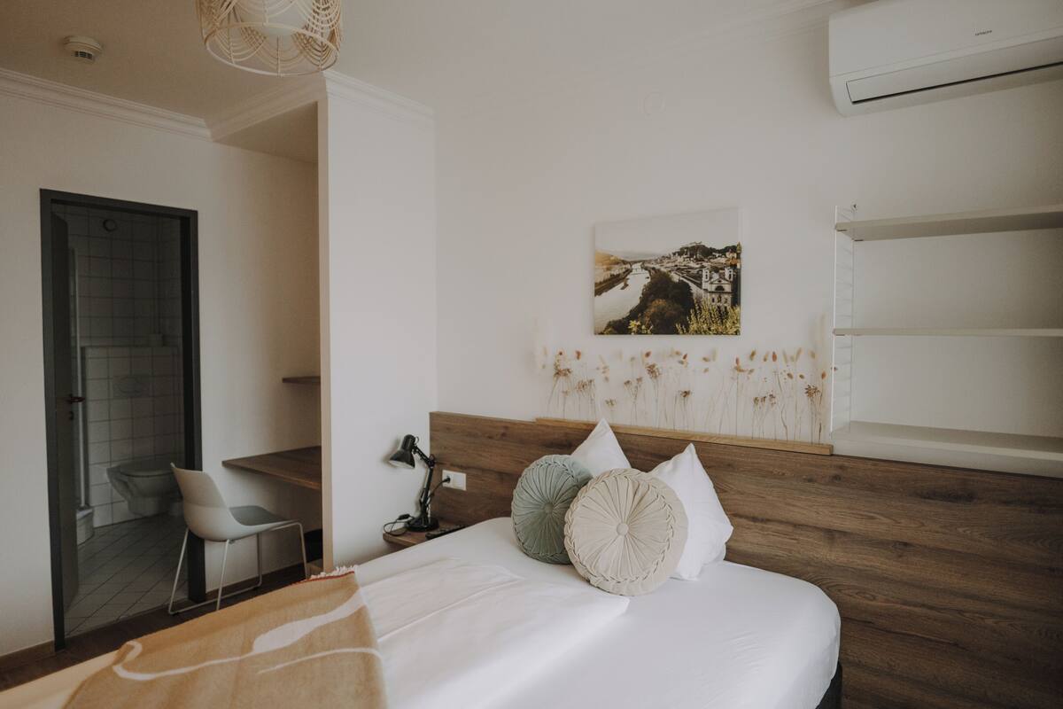 Una habitación de hotel moderna con cabecera de madera, cama con ropa de cama blanca, escritorio con silla, aire acondicionado montado en la pared y un cuadro enmarco con un paisaje.