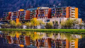 Un hotel de varios pisos con balcones, reflejado en el agua y con montañas al fondo.
