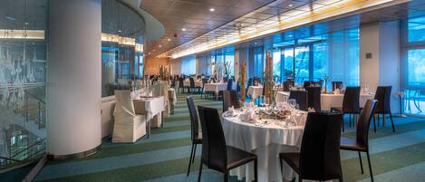 Un comedor amplio con varias mesas y sillas, ventanas grandes que ofrecen vistas al exterior y un diseño de techo moderno.