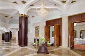 A spacious lobby with a grand chandelier, a round wooden table with floral arrangements, and a large painting on the wall.