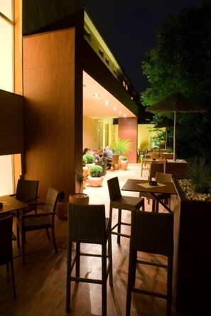 An outdoor dining area with wicker chairs and tables, surrounded by potted plants and a well-lit interior space visible through an open doorway. - Ambient Hotel (Domzale)