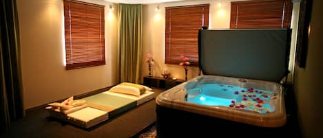 A spa room with a massage table, a hot tub, and two windows with blinds.