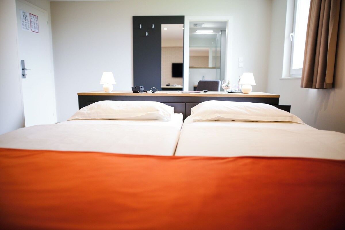 A hotel room with two beds, a desk with a lamp, a mirror, and a window with curtains.