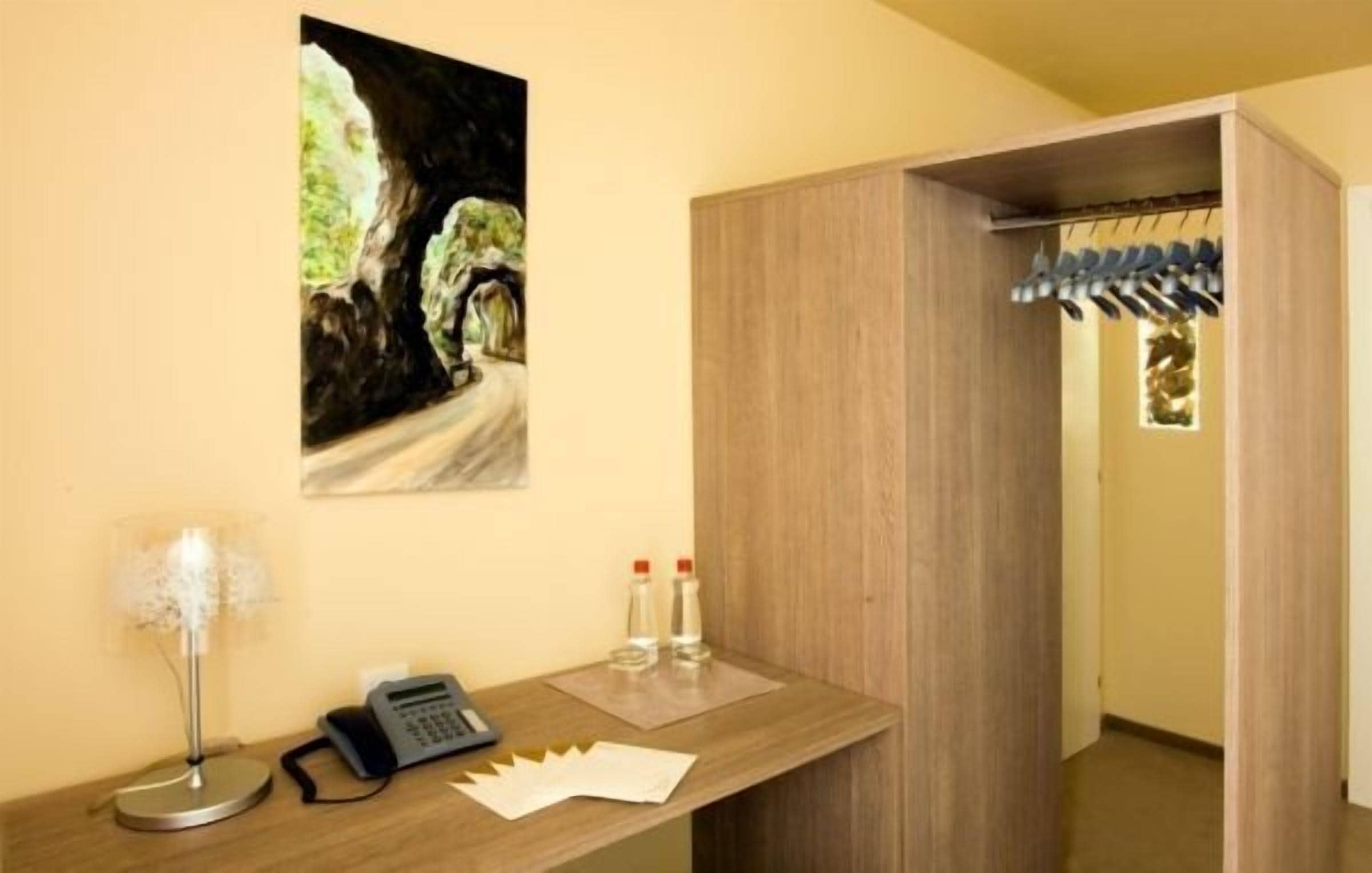 Une chambre d’hôtel avec un bureau en bois, un téléphone, deux bouteilles d’eau, un tableau représentant une grotte et une armoire en bois équipée d’un porte-chaussures suspendu.