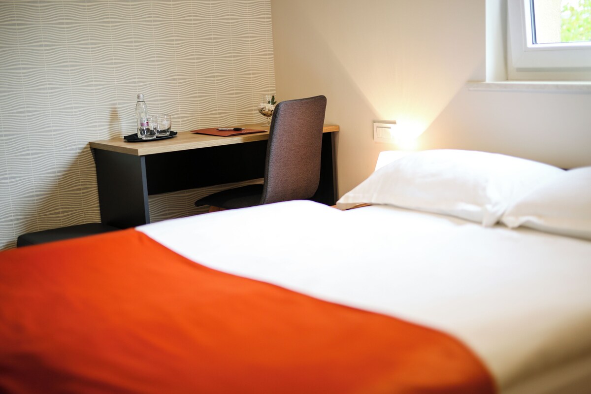 A hotel room with a bed, a desk with a chair, a window, and a patterned wall.
