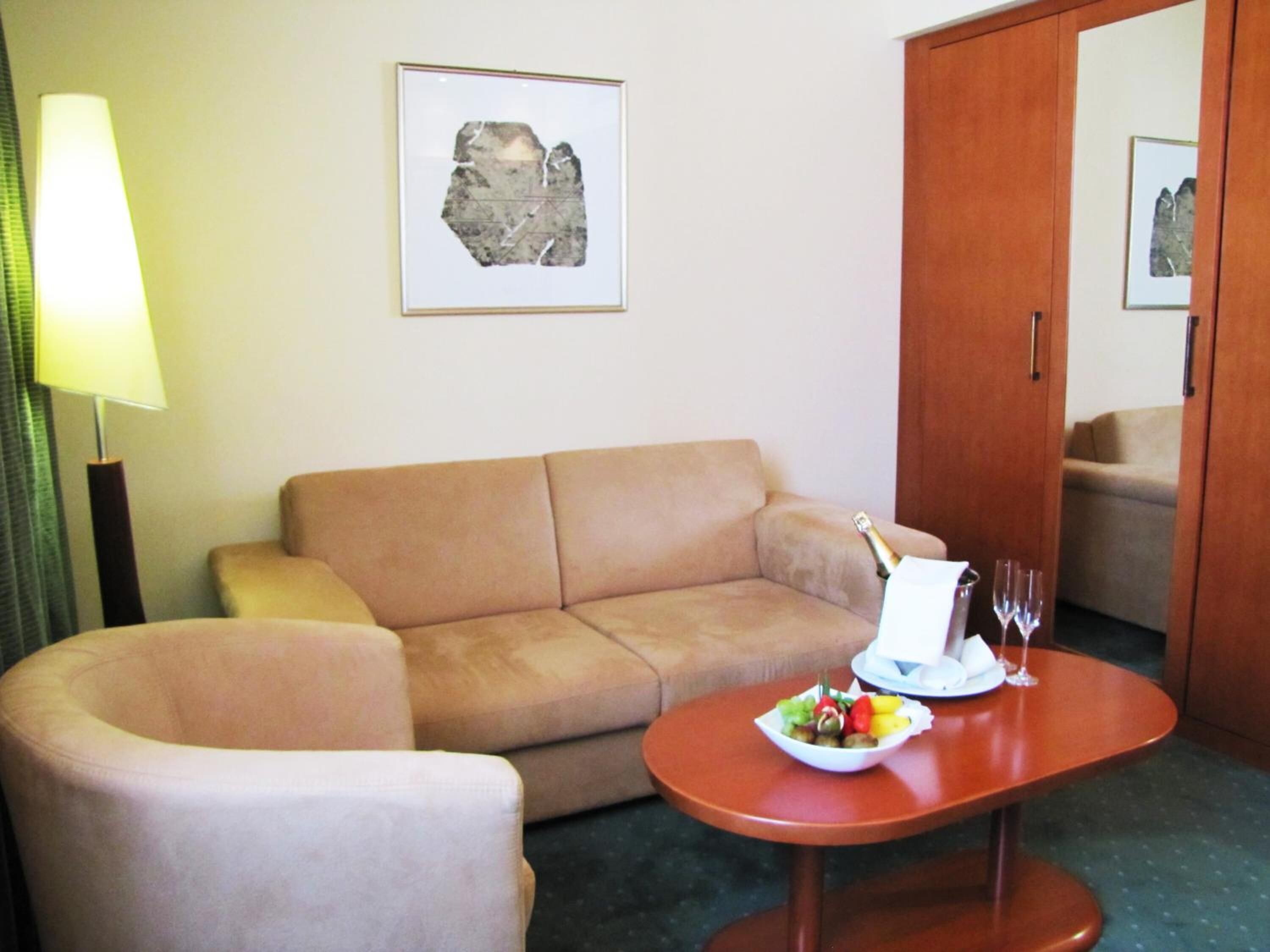 Habitación de hotel con un sofá color beige, una mesita redonda pequeña con un frutero y una botella de champán, una lámpara y un cuadro en la pared.